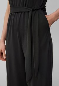Schwarzer Jumpsuit mit einem gebundenen Gürtel in der Taille, aus glattem, strukturiertem Stoff und mit einer Seitentasche. Minimalistisches Design mit klaren Linien.