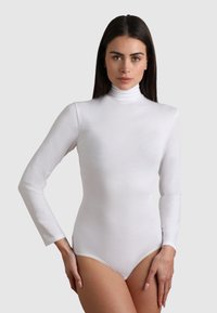 Donna in un body a maniche lunghe bianco con colletto a lupetto, in piedi contro uno sfondo chiaro e uniforme, con i capelli scuri lisci.