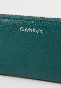 Teal lederen portemonnee met zichtbare textuur, ritssluiting en zilveren "Calvin Klein" logo. Eenvoudig rechthoekig ontwerp.