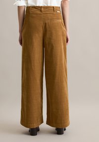 Vrouw in hoge taille, wijde bruine corduroy broek en zwarte hoge hakken, gezien van achteren tegen een effen achtergrond.