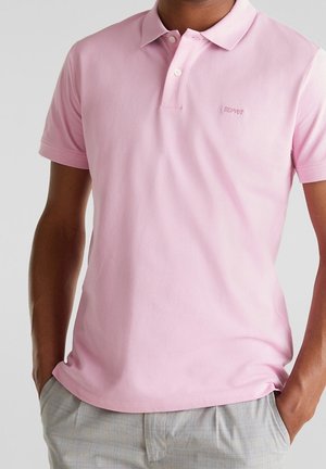 Mand iført lyserød kortærmet polo med krave og knapper, kombineret med lysegrå ternede bukser, hænderne i lommerne.