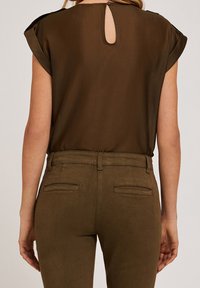 Blusa marrone con maniche corte, tessuto trasparente e dettagli a chiave sul retro. Abbinata a pantaloni verdi oliva aderenti con tasche posteriori.