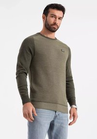 Maglione lavorato a maglia verde oliva con polsini e orlo a costine, presenta una parte anteriore testurizzata e una piccola etichetta con il marchio sul petto.