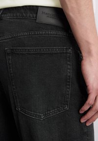 Zbliżenie na tylną kieszeń czarnych dżinsów z widoczną metką marki Marc O'Polo i ręką osoby spoczywającą obok kieszeni.