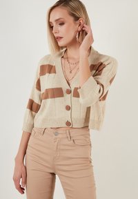Bežový cropped kardigan s hnědými pruhy, V výstřihem, třemi knoflíky a žebrovanými manžetami, spárovaný s bežovými úzkými džíny.