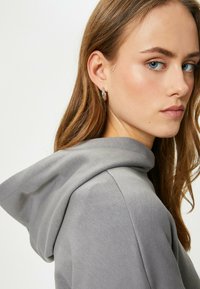 Sudadera gris con una textura suave, que presenta un corte holgado y una gran capucha caída. El modelo tiene el pelo largo y lleva pendientes de aro.