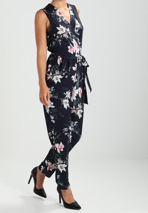 Frau trägt einen ärmellosen schwarzen Jumpsuit mit Blumenmuster und Taillengürtel sowie schwarze High Heels, steht vor einem einfarbigen Hintergrund.