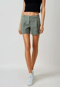 Shorts à carreaux verts et blancs avec un design ajusté, présentant des plis sur le devant et une fermeture à bouton unique. Portés avec un top noir et des baskets blanches.