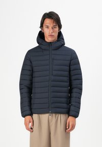 Jonge man in een zwart gewatteerd hooded jack, dichtgeritst, staand met ontspannen armen tegen een effen witte achtergrond.