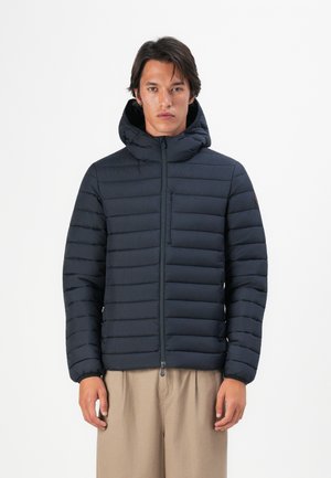 Ung mann iført en svart quiltet hettejacket med glidelås, stående med avslappede armer, mot en enkel hvit bakgrunn.