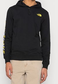 Schwarzer Kapuzenpullover mit gelbem Logo auf der Brust und weißem Schriftzug am Ärmel. Hergestellt aus weichem Stoff, mit Kordelzugkapuze und Taschen.