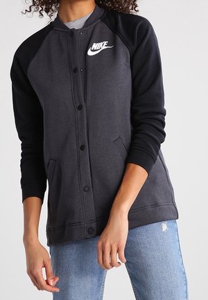 Osoba ubrana w ciemnoszarą i czarną kurtkę Nike na guziki z kieszeniami oraz jasnoniebieskie podarte dżinsy, stojąca na jednolitym tle.