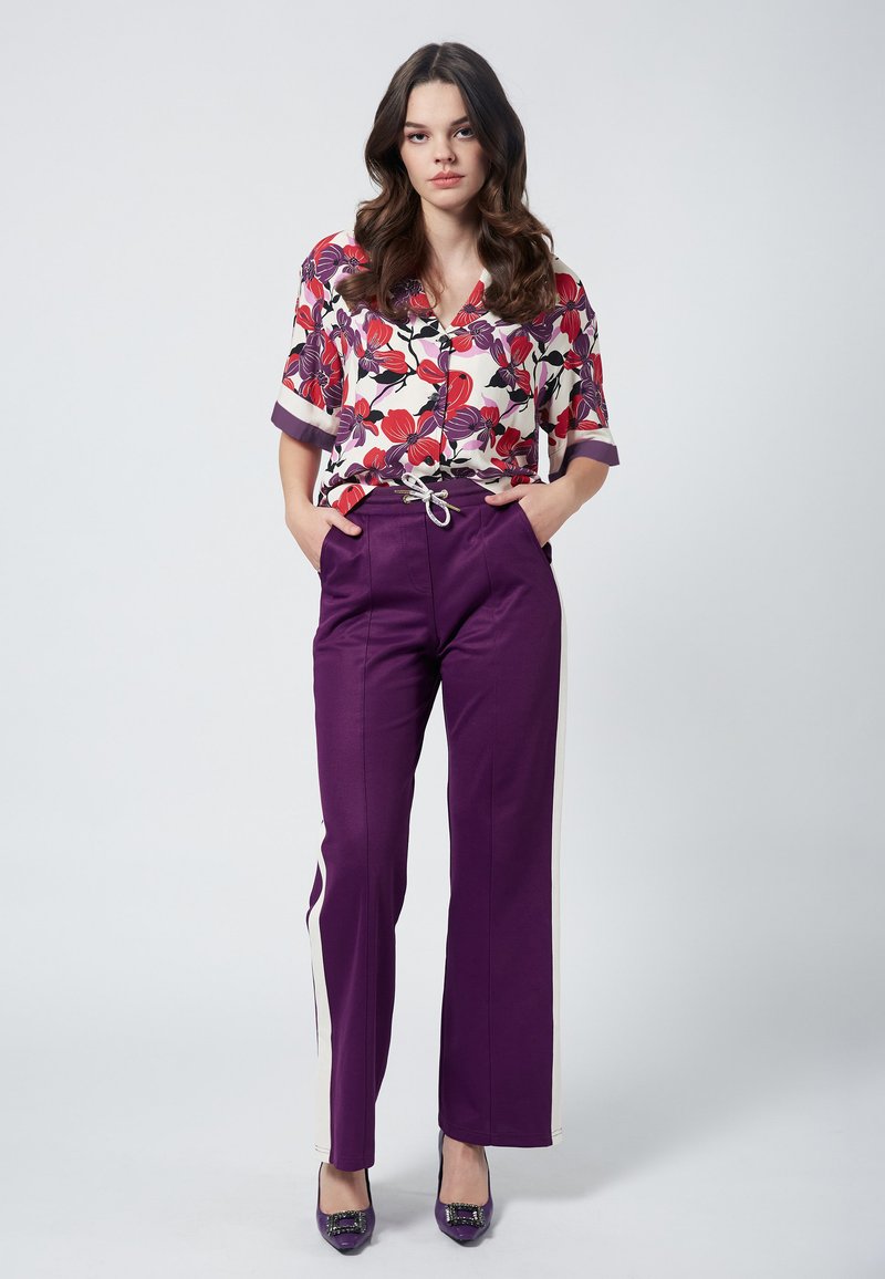 Koszula w kwiaty na guziki w kolorze białym, czerwonym i fioletowym; luźny krój. Fioletowe spodnie proste z regulowanym paskiem i kremowymi paskami po bokach.