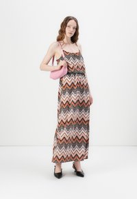 Maxi-jurk met een veelkleurig zigzagpatroon in oranje, zwart en wit; verstelbare bandjes; gecombineerd met een kleine roze handtas. Zwarte puntige hakken.
