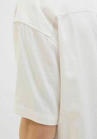 T-shirt med korte ærmer i lyse beige stof, der har en glat tekstur og et enkelt design med en rund halsudskæring og syede sømme.