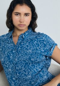 Blå bluse med hvitt blomsterdesign, korte ermer og krage. Mykt, lett stoff med knappdetalj.