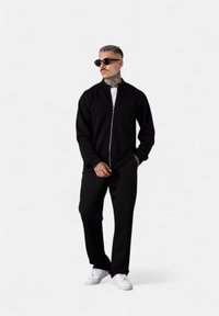 Schwarze Zip-up-Jacke mit hohem Kragen, kombiniert mit passenden schwarzen Hosen und weißen Sneakers. Die Accessoires umfassen dunkle Sonnenbrillen.