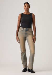 Γυναίκα με σκούρα μαλλιά, φορώντας μαύρο αμάνικο τοπ, ξεβαμτζέ μπεζ τζιν και μαύρα μποτάκια, στέκεται μπροστά σε απλό φωτεινό φόντο.