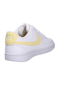 Scarpa Nike bianca con una morbida texture in pelle, caratterizzata da uno swoosh giallo e una linguetta sul tallone. La suola è bianca con leggere macchie.