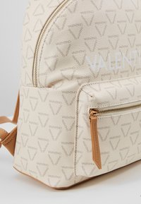 Sac à dos en cuir beige avec un motif logo sur toute sa surface, forme arrondie, poche avant zippée et accents beige sur la tirette et les sangles.
