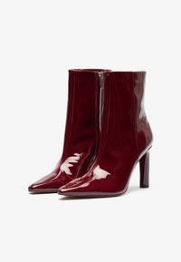 Botines de charol burdeos con puntas afiladas, una textura suave, cremallera lateral y un tacón alto delgado.
