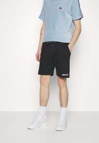 Chemise pour hommes en velours côtelé bleu clair à manches courtes, col rond, avec un petit logo orange. Portée avec un short noir à cordon de serrage et des baskets blanches.