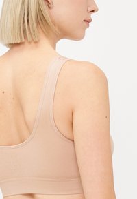 Beige naadloze bh met brede bandjes, gestructureerde ribbelstof en een eenvoudige haaksluiting op de schouder, die een strakke, minimalistische uitstraling toont.