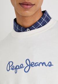 Sudadera blanca con el logo de "Pepe Jeans" bordado en azul, llevada sobre una camisa de cuadro azul marino con cuello y detalles de botón, que presenta texturas contrastantes.