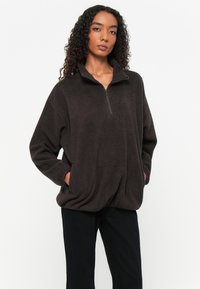 Donkerbruin fleece pullover met een halvemaan-rits kraag, lange mouwen en zijzakken. Zachte textuur en ontspannen pasvorm voor comfort.