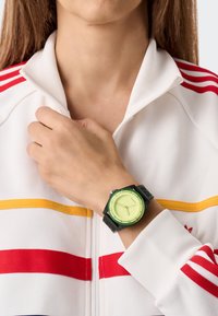 Roheline ja kollane käekell valge lukuga jakil, millel on punased ja kollased triibud. Kummist rihm ja ümar nägu.