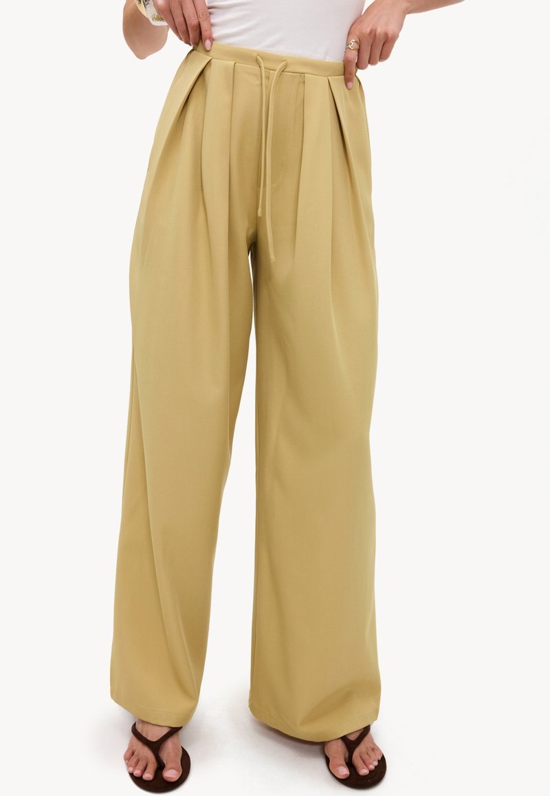 Personne portant un pantalon beige ample taille haute avec des plis devant et un cordon, assorti à des sandales marron foncé et un haut blanc.