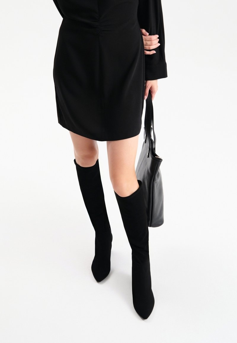 Femme portant une mini-robe noire à manches longues, des bottes noires montantes à bout pointu, tenant un sac à main en cuir noir à ses côtés.
