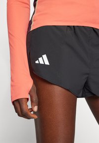 Sorte træningsshorts med et hvidt trekantet logo på venstre side, parret med en ferskenfarvet langærmet skjorte. Har et slankt, tætsiddende design.