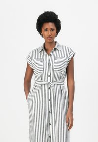 Rochie fără mâneci, cu nasturi, de culoare albă cu dungi verticale negre, cu două buzunare la piept, curea la talie și textură de țesătură netedă.