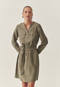 Olivgrünes Kleid mit langen Ärmeln, Knopfkragen, Brusttaschen und einem Taillengürtel. Glatte Stofftextur.