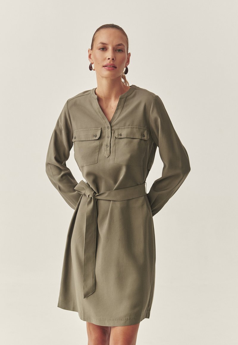 Olivgrünes Kleid mit langen Ärmeln, Knopfkragen, Brusttaschen und einem Taillengürtel. Glatte Stofftextur.