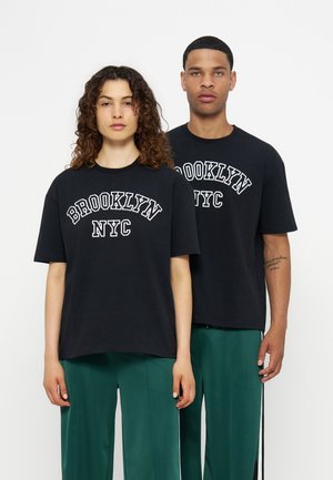 To voksne, der står med fronten mod kameraet, iført matchende sorte "Brooklyn NYC"-t-shirts og grønne bukser mod en ensfarvet hvid baggrund.