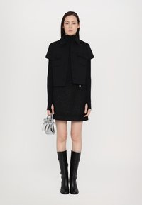 Gilet noir court avec poches à rabat, col roulé à manches longues, jupe noire texturée, bottes noires arrivant au genou et un petit sac à main en argent.