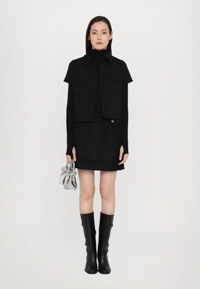 Black cropped vest with flap pockets, long-sleeve turtleneck, textured black skirt, knee-high black boots, and a small silver handbag.