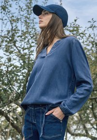 Femme portant une casquette bleue, un haut à manches longues bleu et un jean, debout en plein air, une main dans la poche, regardant sur le côté avec un fond d'arbres.