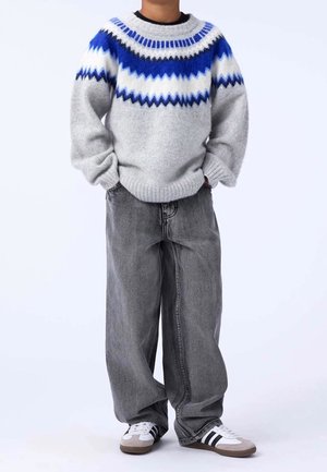 Barn iført en grå sweater med blå og hvide mønstre, løse mørkegrå jeans og hvide sneakers med sorte striber, stående med hænderne i lommerne.