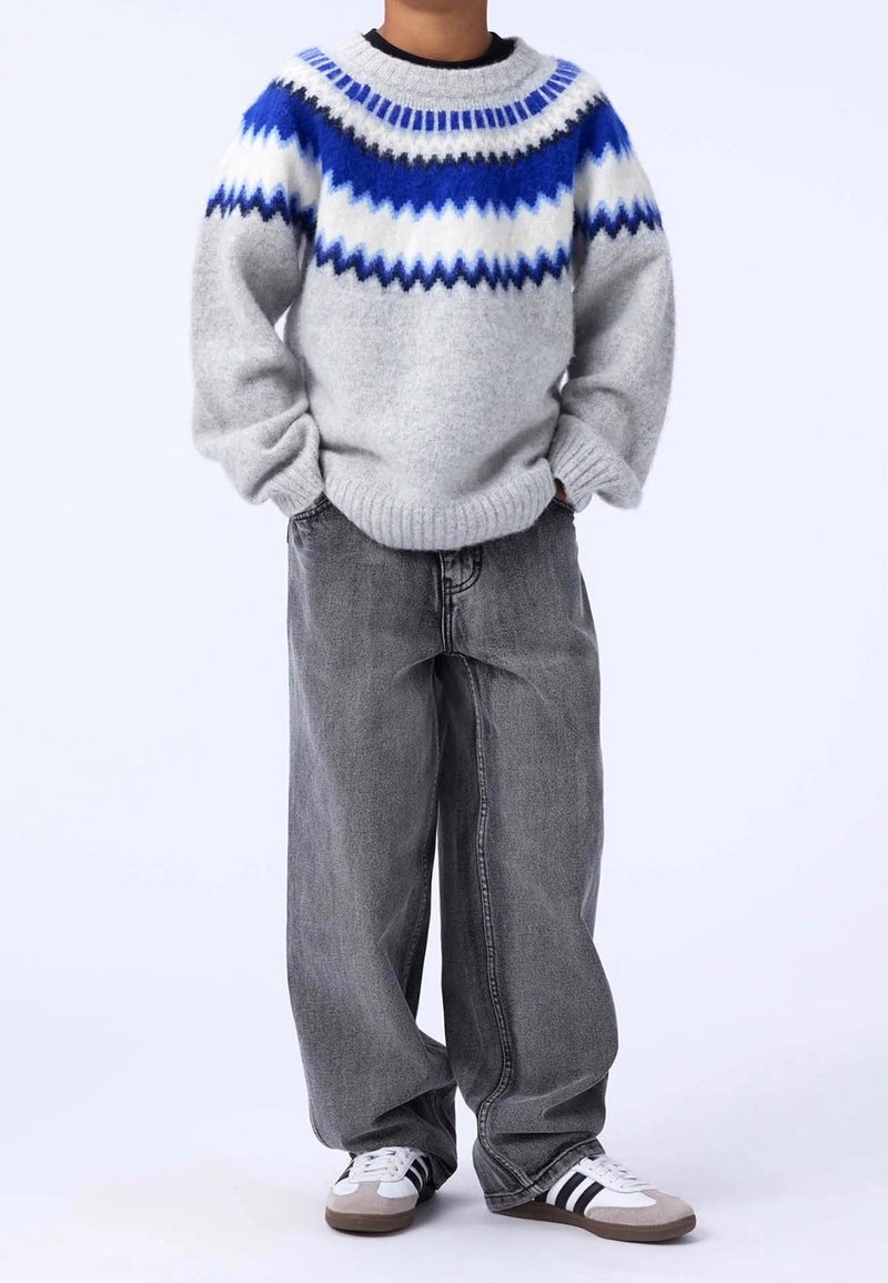 Enfant portant un pull gris avec des motifs bleus et blancs, un jean large gris foncé et des baskets blanches avec des rayures noires, debout les mains dans les poches.