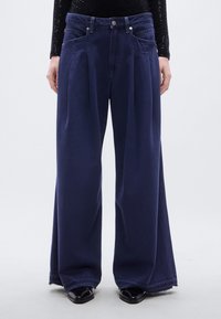 Μακριά παντελόνια σε σκούρο μπλε ντένιμ, με πιέτες μπροστά, σχέδιο με πέντε τσέπες και ξεφτισμένα τελειώματα, σε συνδυασμό με μαύρα παπούτσια.