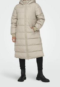 Lange, beige pufferjas met capuchon, voorzien van een ritssluiting aan de voorkant, zijzakken, geribbelde boorden en een horizontaal doorgestikt patroon.