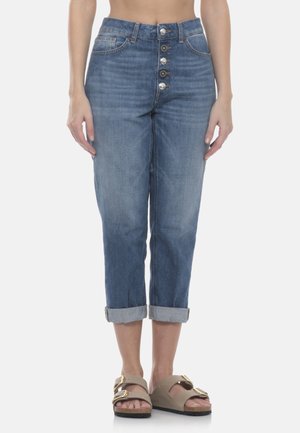 Donna che indossa jeans blu a vita alta, risvoltati, con patta a bottoni a vista e sandali beige con fibbie dorate.