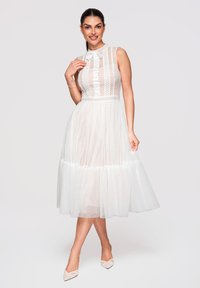 Femme en robe midi blanche en dentelle sans manches avec col à nœud, debout avec une main près du décolleté et portant des chaussures blanches pointues.