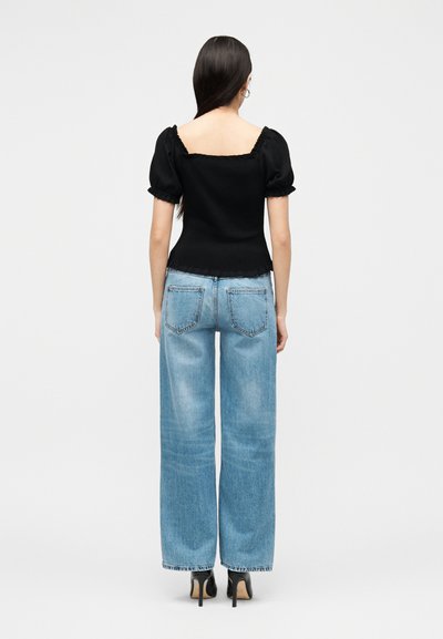 Γυναίκα που στέκεται με την πλάτη, φοράει μαύρο τοπ με φουσκωτά μανίκια, ανοιχτό μπλε τζιν παντελόνι με φαρδιά μπατζάκια και μαύρες ψηλοτάκουνες γόβες πάνω σε λευκό φόντο.