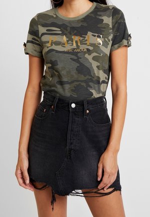 Kvinde iført grøn camouflage T-shirt med teksten "PARIS AVEC AMOUR" i guld og sort distressed denim mininederdel, stående mod en grå baggrund.
