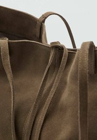 Sac fourre-tout en daim marron, avec de longues poignées, des coutures visibles, et un design non structuré avec une ouverture sur le dessus. Pas de matériel ni d'ornements.