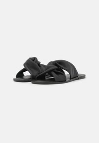 Svarta platta sandaler med en knuten design av mjukt material. Sulan är minimal och platt, vilket ger ett enkelt och öppet tåutseende.